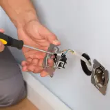 Ręce majsterkowicza przykręcają śrubokrętem mechanizm gniazdka. Widać przewody elektryczne i puszkę instalacyjną.
