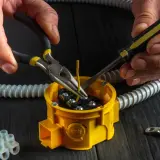 Ręce elektryka podczas osadzania puszek elektrycznych, używając szczypiec i śrubokręta do podłączenia przewodów.