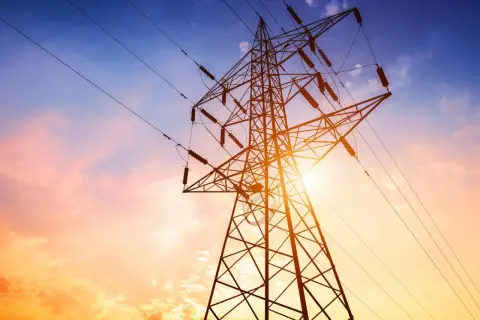 Słup energetyczny na działce, zasilający dom. Zachód słońca maluje niebo na pomarańczowo i niebiesko.