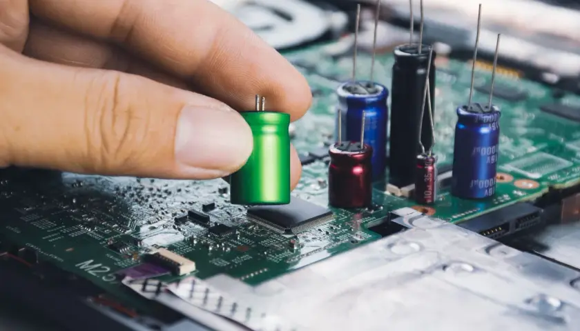 Dłoń trzyma zielony kondensator, przygotowując go do montażu na płycie głównej. W tle widać inne kondensatory i układy elektroniczne.