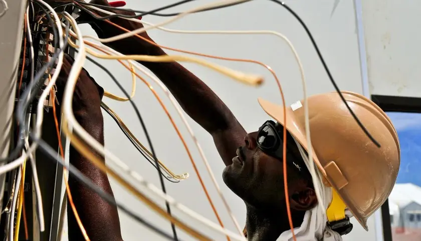 Elektryk w kasku i okularach pracuje przy skrzynce z przewodami. Nauka jak zostać elektrykiem wymaga precyzji.