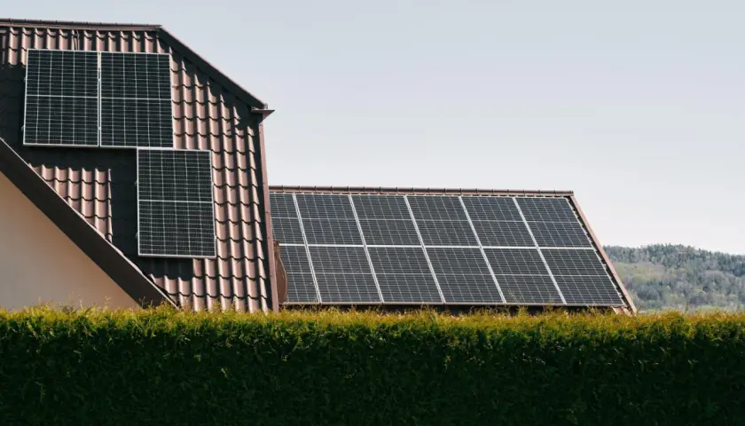 Panele słoneczne na dachu domu, zapewniające czystą energię. W tle las, a na pierwszym planie żywopłot. System jest też zabezpieczony odgromnikiem.