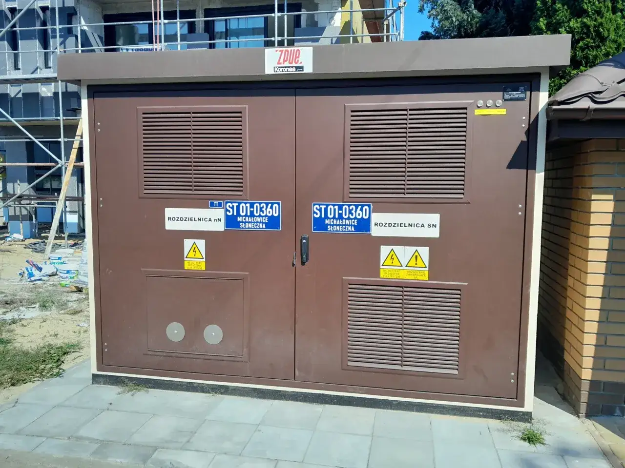 Brązowa stacja transformatorowa z kratkami wentylacyjnymi i tabliczkami informującymi o dystrybucji energii elektrycznej.